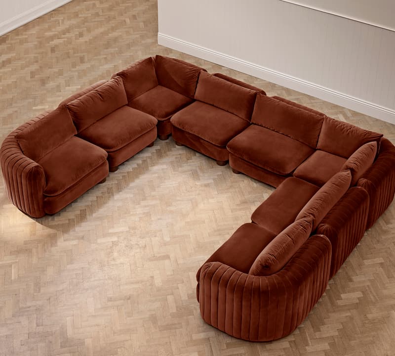 A large, brown, U-shaped sectional sofa is situated on a light wooden parquet floor in a neutral-colored room with white paneled walls.