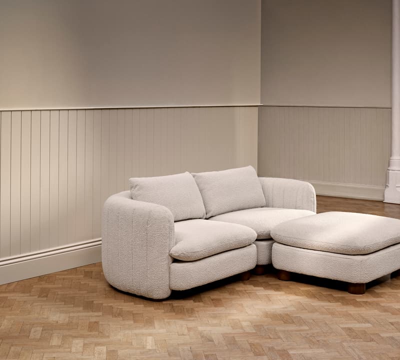 A light beige upholstered sectional sofa with an ottoman sits on a parquet wooden floor against a wall with vertical wainscoting in a softly lit room.