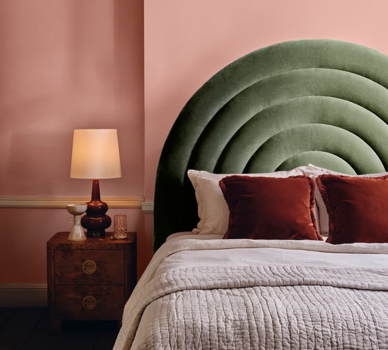 Bed with arched green headboard adorned with two rust-colored pillows in a brightly lit bedroom; beside it, a wooden nightstand with a red lamp, decor items, and an artwork leaning against the wall.