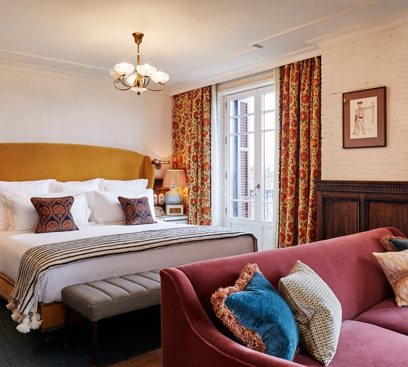 A large bed with a mustard-colored headboard is arranged with white linens and patterned pillows, facing a mauve sofa. The room features floral curtains, a chandelier, and framed artwork.
