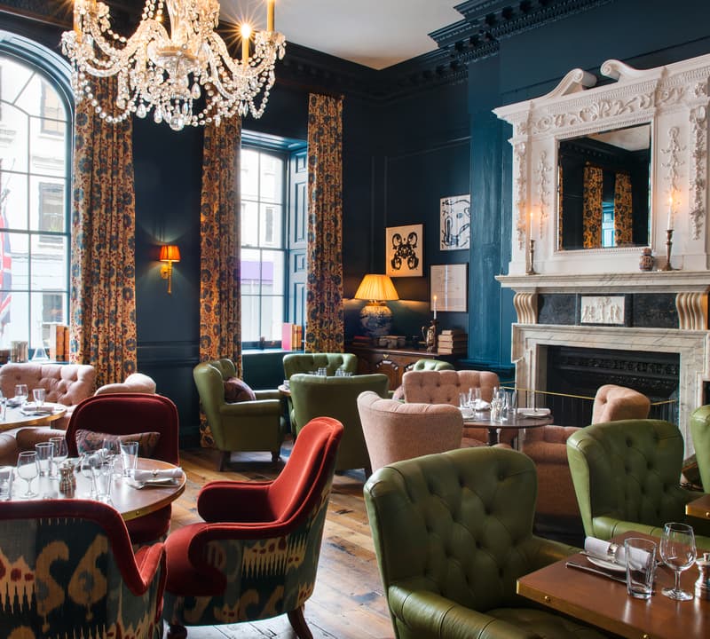 A room with elegant, colorful furniture, a large chandelier, and set dining tables; ornate dark walls and large windows with patterned curtains frame the opulent interior.