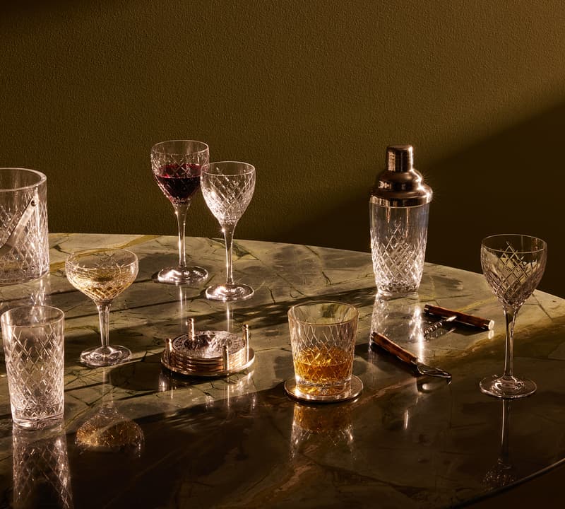 Various crystal glassware, including wine glasses and a cocktail shaker, rest on a marble table under warm light, alongside an ice bucket, coasters, and a corkscrew.