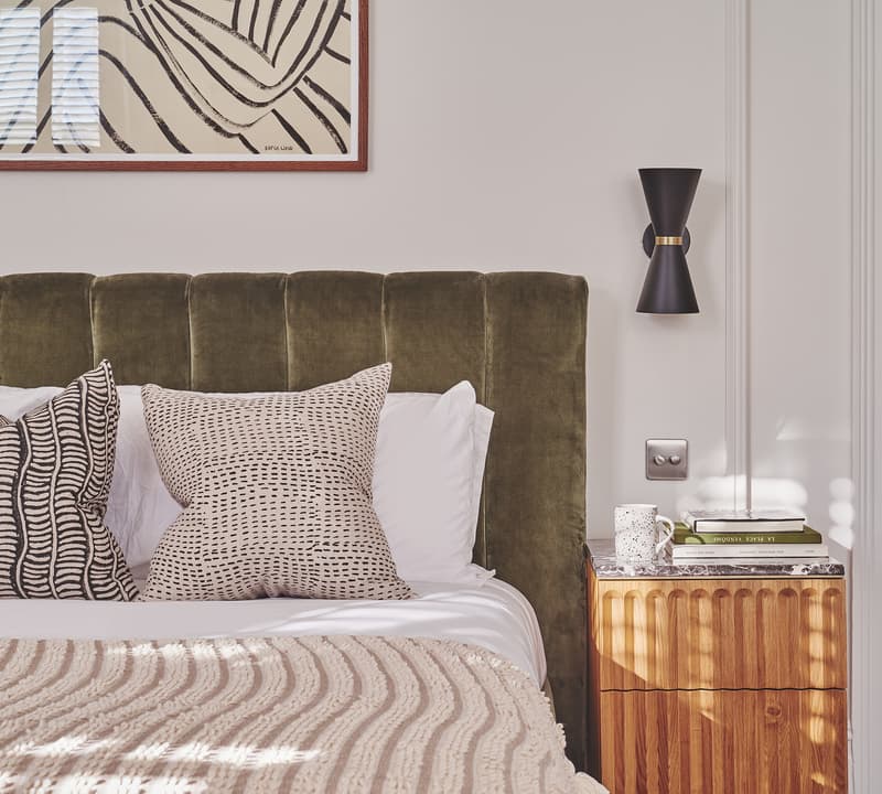 A bed with a green upholstered headboard is adorned with patterned pillows and a knit blanket. A wooden nightstand holding books and a mug sits beside the bed, beneath a wall sconce and abstract artwork.