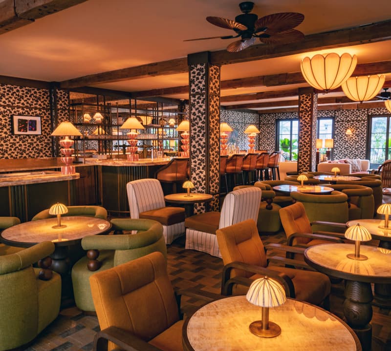 Comfortable lounge chairs and tables with small lamps fill an elegant, warmly lit restaurant-bar with patterned wallpaper, wooden beams, and large windows showcasing greenery outside.