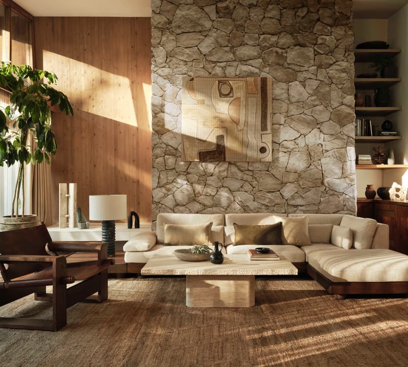 A beige sectional sofa and a wooden armchair sit in a cozy living room with a stone wall, large plant, abstract art, and wooden shelves filled with decor items.