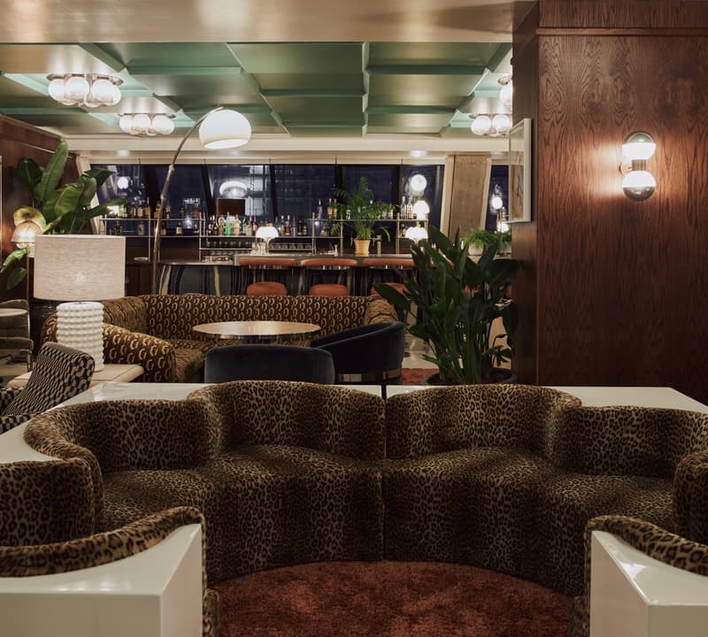 A leopard print sofa sits in a stylish lounge area with plants, tables, and lamps, against the backdrop of a bar with stools and various bottles in the background.