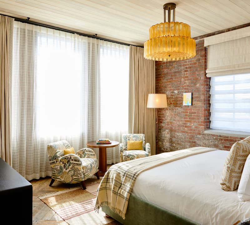 A cozy bedroom with a large bed, two armchairs beside a round wooden table, all illuminated by natural light from floor-to-ceiling windows and a golden chandelier. Brick walls frame the space.