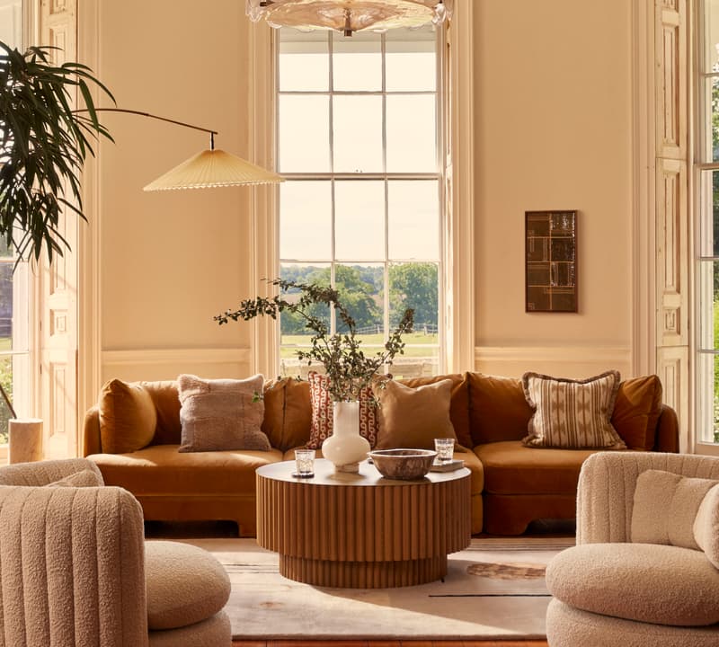 A cozy living room features a mustard-colored velvet sectional adorned with pillows, a round wooden coffee table with decor, plush armchairs, and large windows offering a scenic view outside.