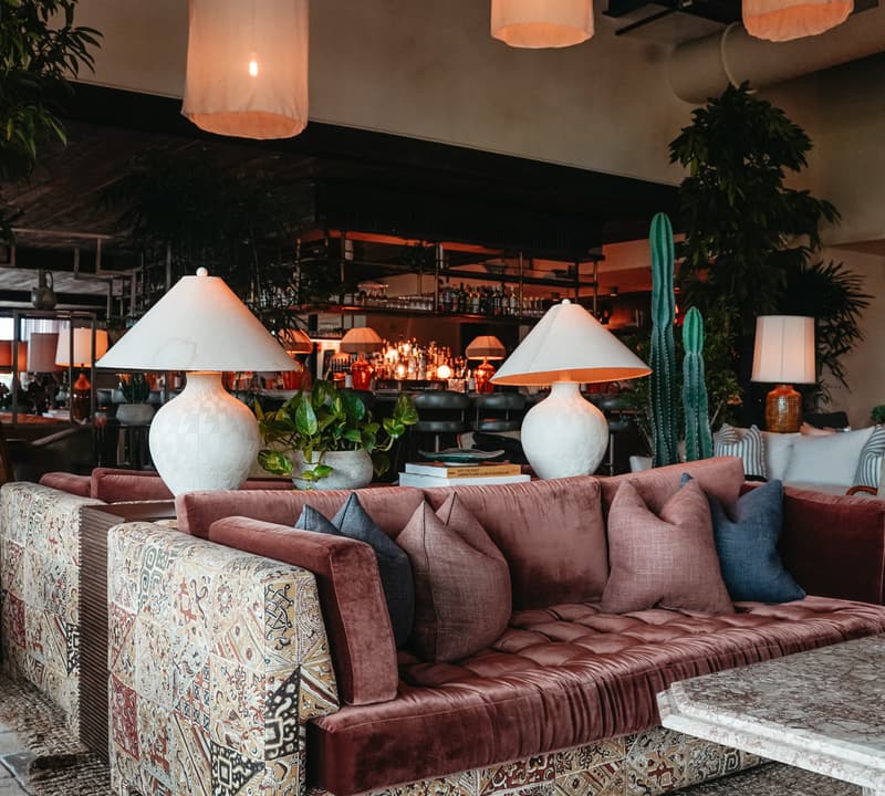 A patterned, plush sofa adorned with cushions and large table lamps stands in a cozy, dimly-lit lounge with plants and pendant lights, adjacent to a bar area.