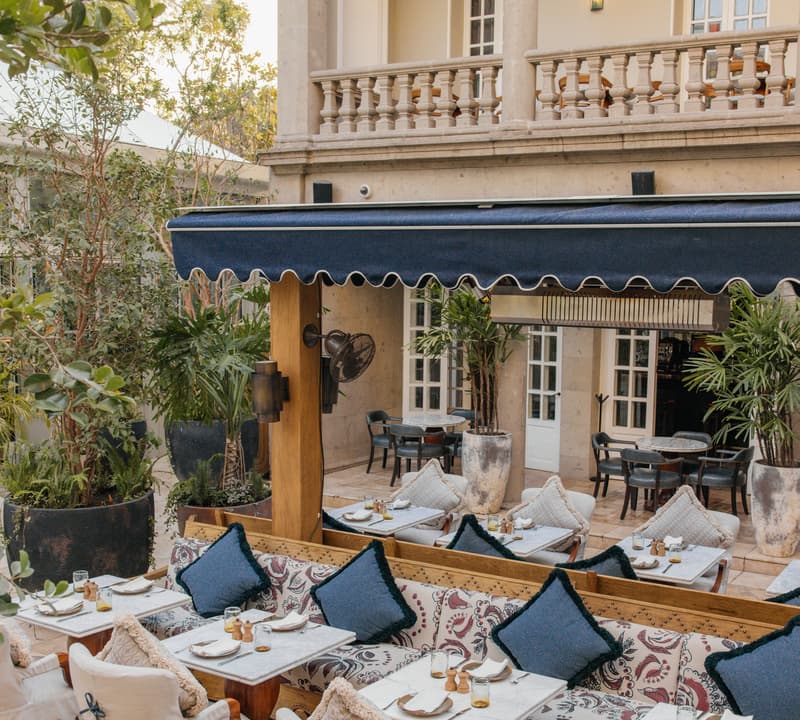 Outdoor dining area with cushioned chairs and blue pillows arranged around tables beneath a blue awning. Surrounded by potted plants and overlooked by a beige, columned building with balconies.