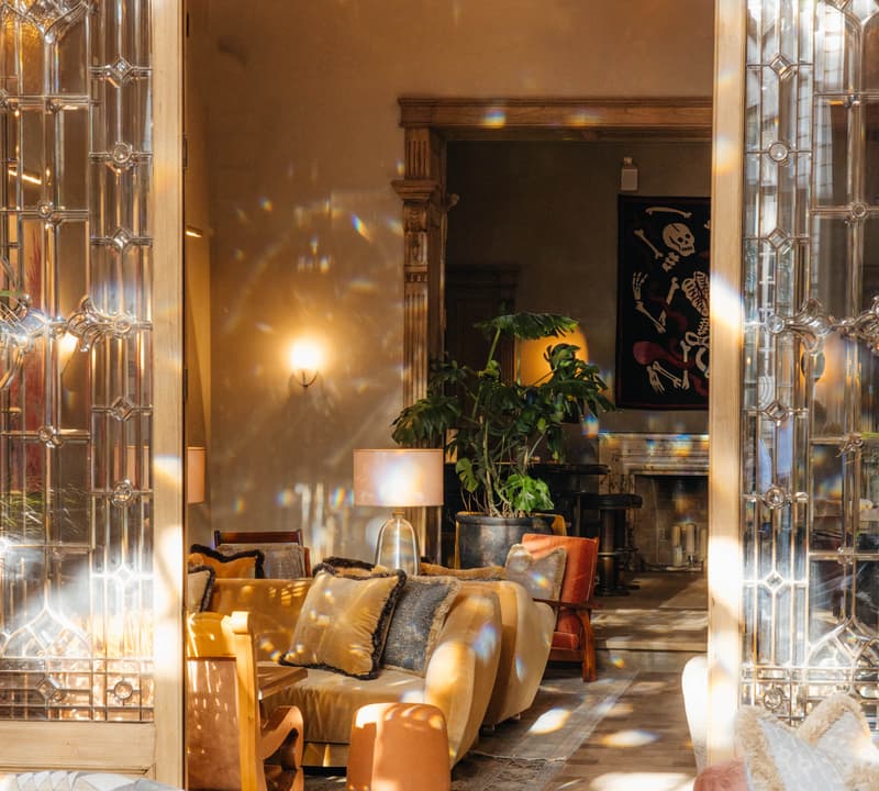 Luxurious living room features plush sofas, ornate lamps, and lush plants. Elegant stained-glass doors and chandeliers enhance the sophisticated ambiance. Sunlight filters through the room, creating a warm, inviting atmosphere.
