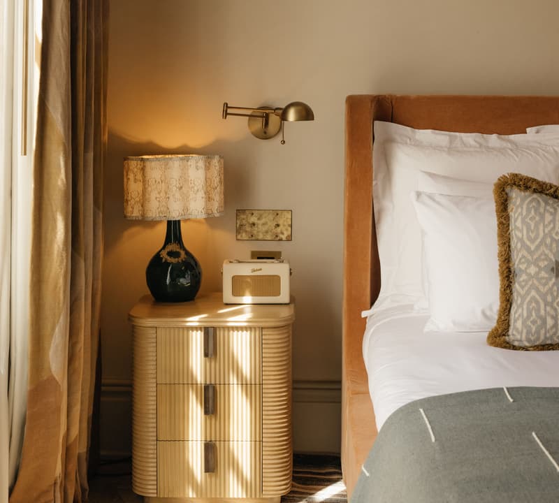 A wooden nightstand with a lamp, a small beige radio, stands beside a neatly made bed with white linens and a gray blanket, in a warmly lit bedroom. Shadows from window blinds fall across the floor.