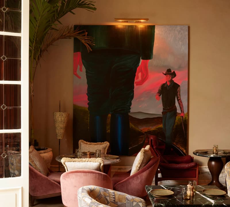 Large painting of a figure in a hat dominates a cozy, tastefully-decorated café with plush chairs, green marble tables, and warm lighting. Palm fronds and open French doors add elegance.