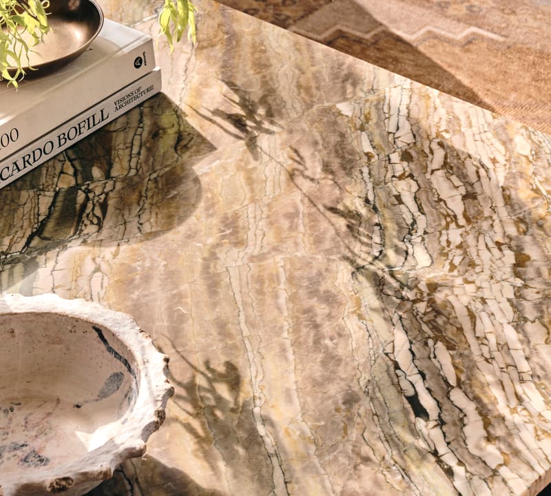 Marble tabletop with a rustic bowl, a book titled “Ricardo Bofill” beneath a small bronze dish, under dappled sunlight with hanging green leaves, situated on a patterned rug.