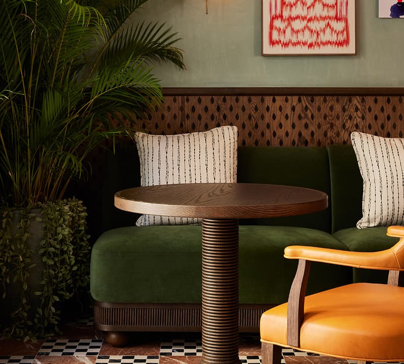 A wooden round table stands between a green cushioned bench and a tan leather chair, set against a wall with modern artworks and indoor plants.