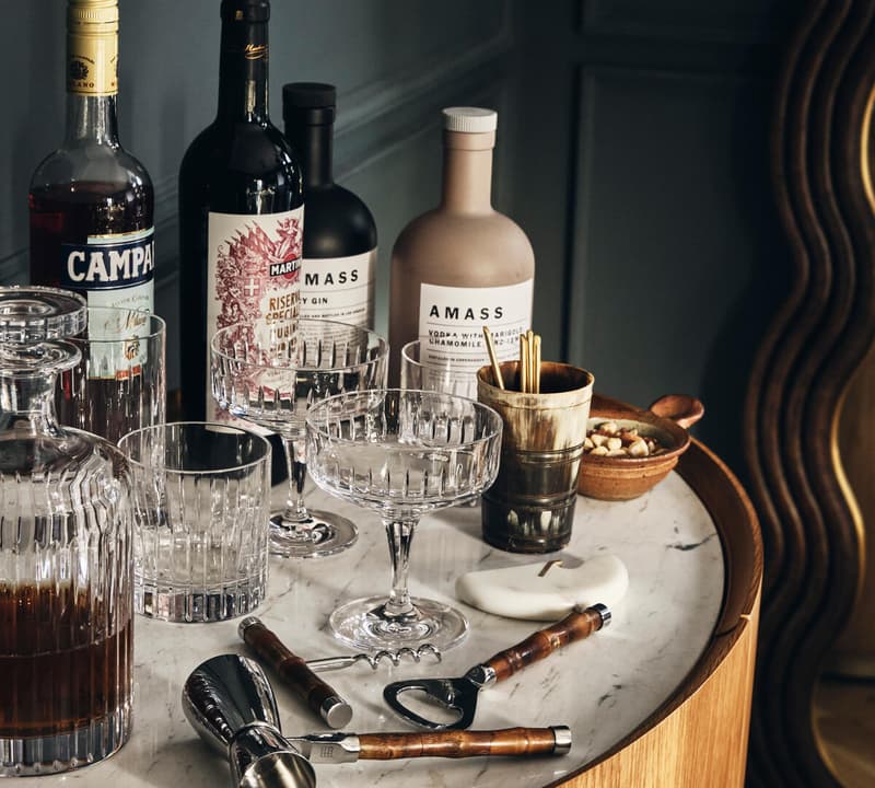 Bottles and glasses arranged on a marble-topped wooden cabinet with cocktail tools, peanuts in a small bowl, and a stylized mirror reflecting part of the dimly lit room. Text includes: "CAMPARI," "MARTINI," and "AMASS."