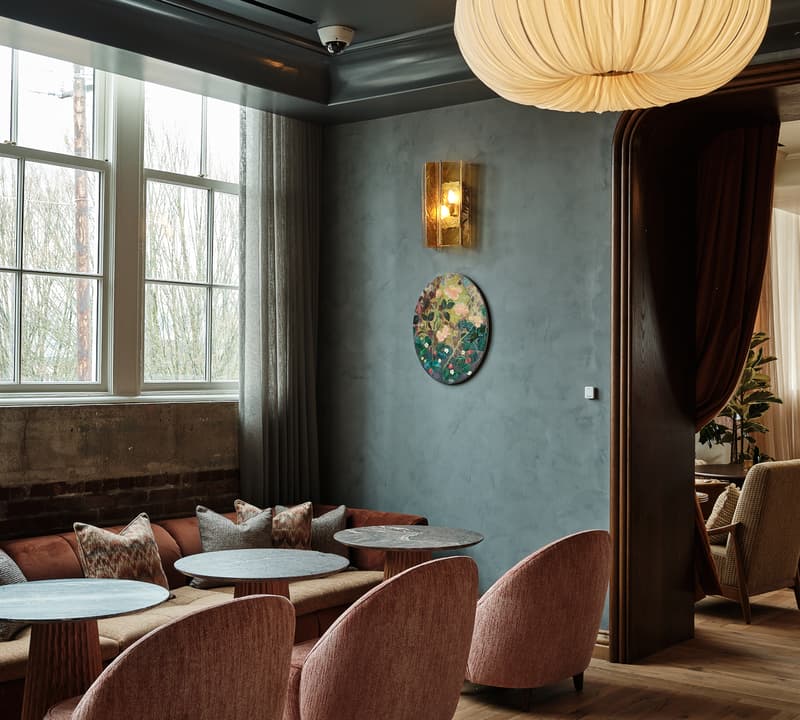 A cluster of round tables and pink upholstered chairs arranged around a corner seating area with large windows; above, a beige pendant light and wall art adorn an elegantly furnished room.