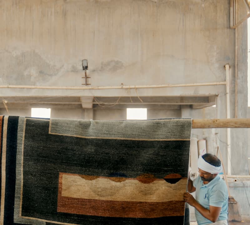 A person in traditional attire is hanging a large, patterned carpet over a wooden beam in an industrial building with a corrugated metal roof and birds perched above.
