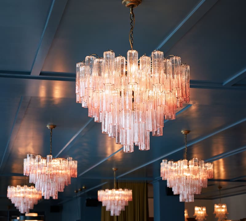 Multiple glass chandeliers hang from a ceiling, illuminating a spacious room with a blue ceiling and walls, and additional chandeliers visible in the background.