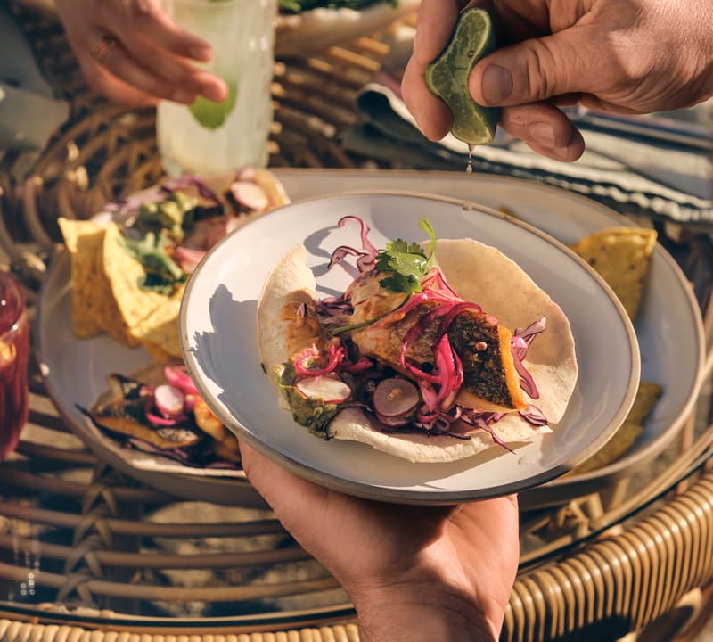 Plate with taco held by a hand, lime squeezed above it; tacos, chips, drinks, and salads on wicker table outdoors.