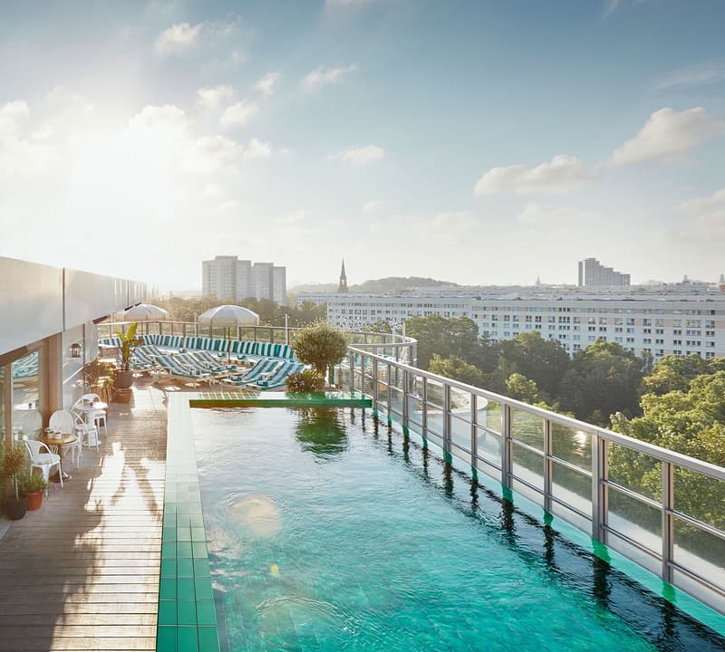 Rooftop infinity pool reflecting sunlight, flanked by lounge chairs and potted plants, with city buildings and greenery visible in the background.