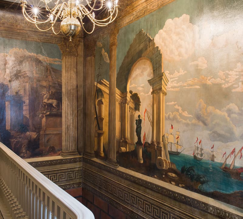 A mural featuring ancient ruins and ships at sea adorns the walls of an elegant staircase, illuminated by a grand chandelier.