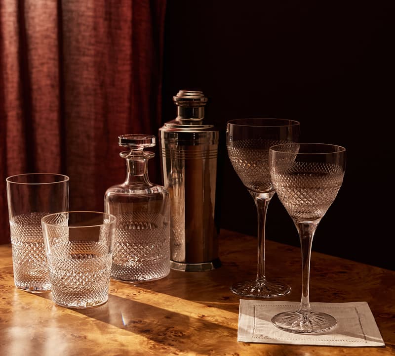 Crystal glasses and a decanter stand on a wooden table next to a cocktail shaker, bathed in warm light from a nearby window with red curtains.