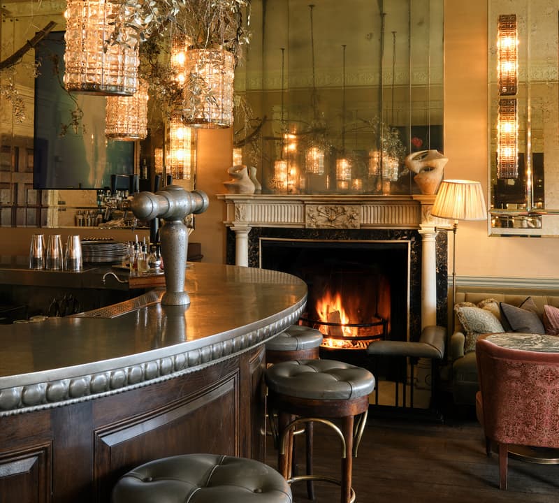 A curved, wooden bar with cushioned stools sits in front of a lit fireplace surrounded by seating. Warm lighting comes from elegant, vintage-style chandeliers, creating a cozy ambience.