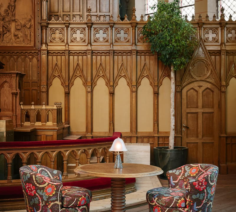 Two patterned chairs are positioned around a round wooden table with a small lamp, set within a richly detailed wooden church interior, complete with organ pipes, stained glass windows, and red cushioned seats.