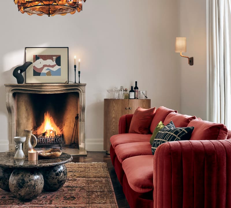 A red sofa sits in front of a lit fireplace surrounded by a rug, coffee table, artwork, and wine bottles, under a decorative chandelier in a cozy living room.
