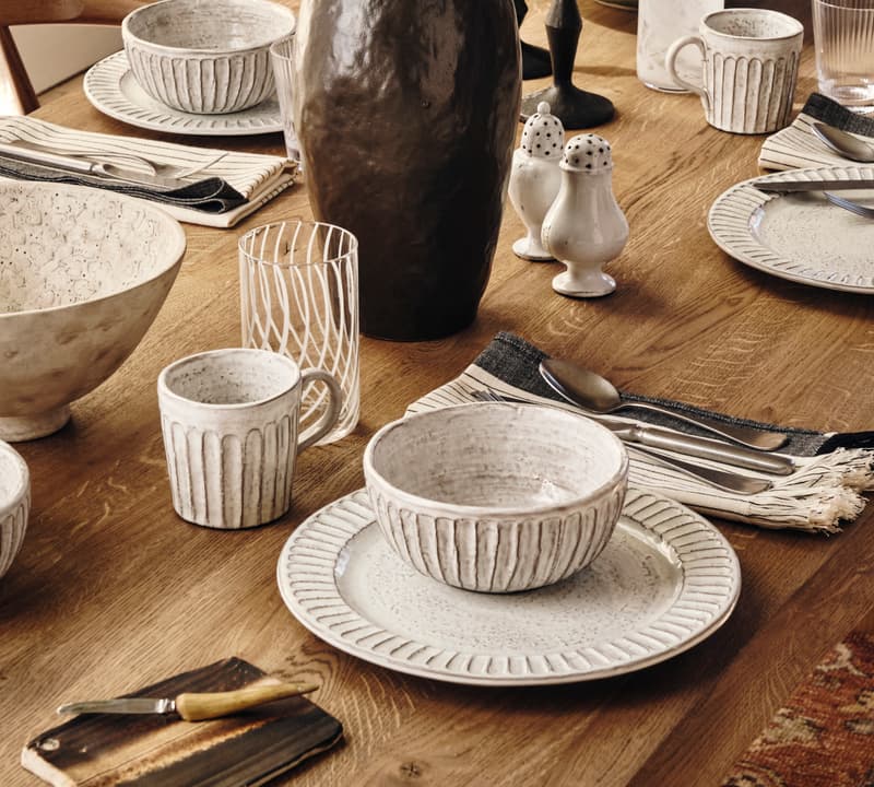 A wooden dining table is set with white ceramic dishes and mugs, a brown vase with green leaves, cutlery, napkins, a bowl of apples, and a salt and pepper shaker.