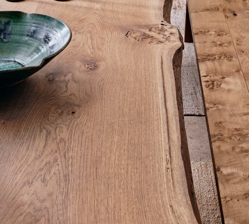 A natural wood table with a wavy edge holds a green ceramic bowl and three black candle holders, set in a rustic room with a matching wooden bench and a textured rug.