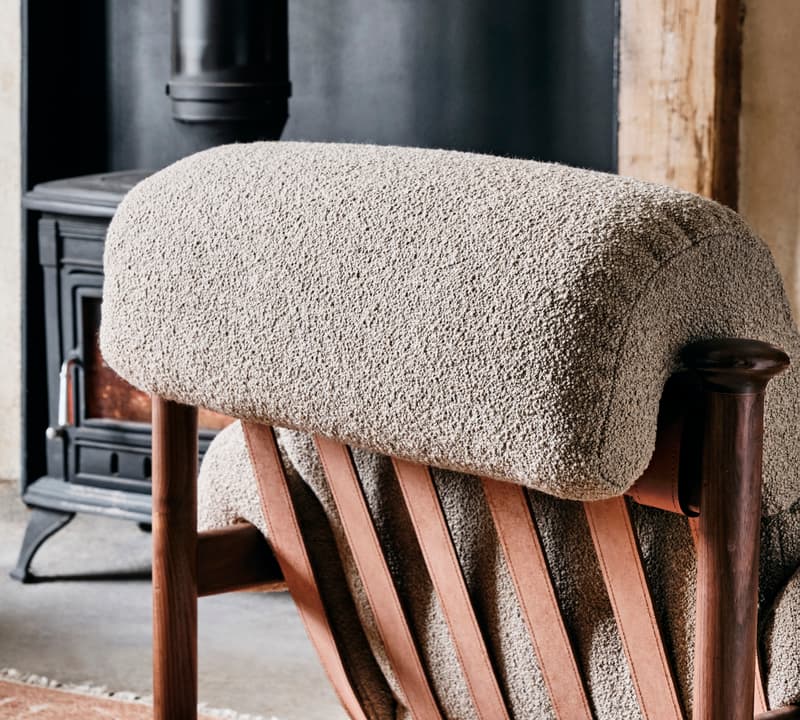 A cozy, high-backed lounge chair with cushioned upholstery and wooden frame faces a black wood-burning stove in a warmly lit, rustic room with wooden beams.