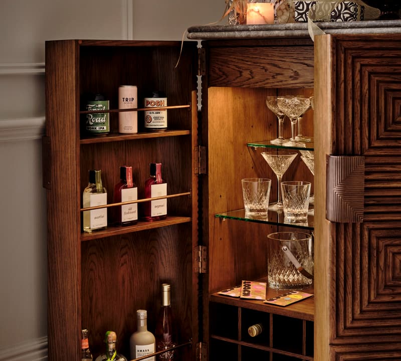 A wooden liquor cabinet stands open revealing various bottles, glasses, and barware. Above the cabinet, candles and a bottle are arranged decoratively. The setting is a warmly-lit corner of a room.
