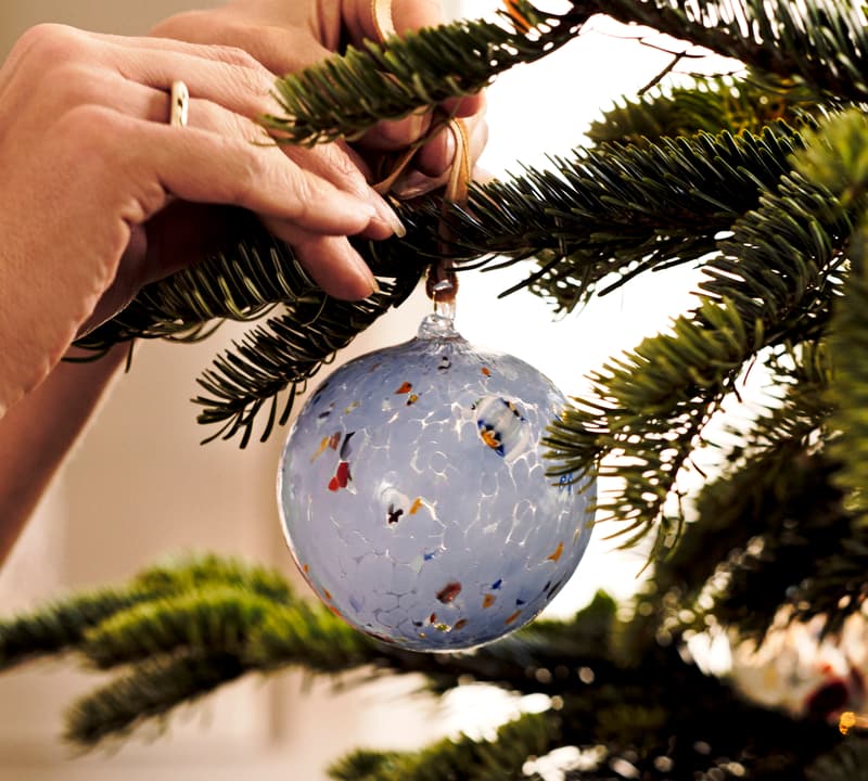 A hand carefully hangs a blue glass ornament with colorful specks on a green Christmas tree inside a warmly lit room.