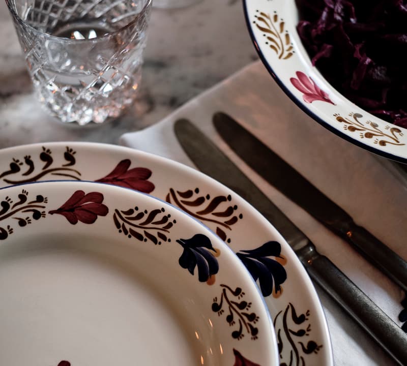 Patterned ceramic plates stacked together, a glass of water, and cutlery on a white cloth napkin, placed on a marble table beside another plate containing dark red food.