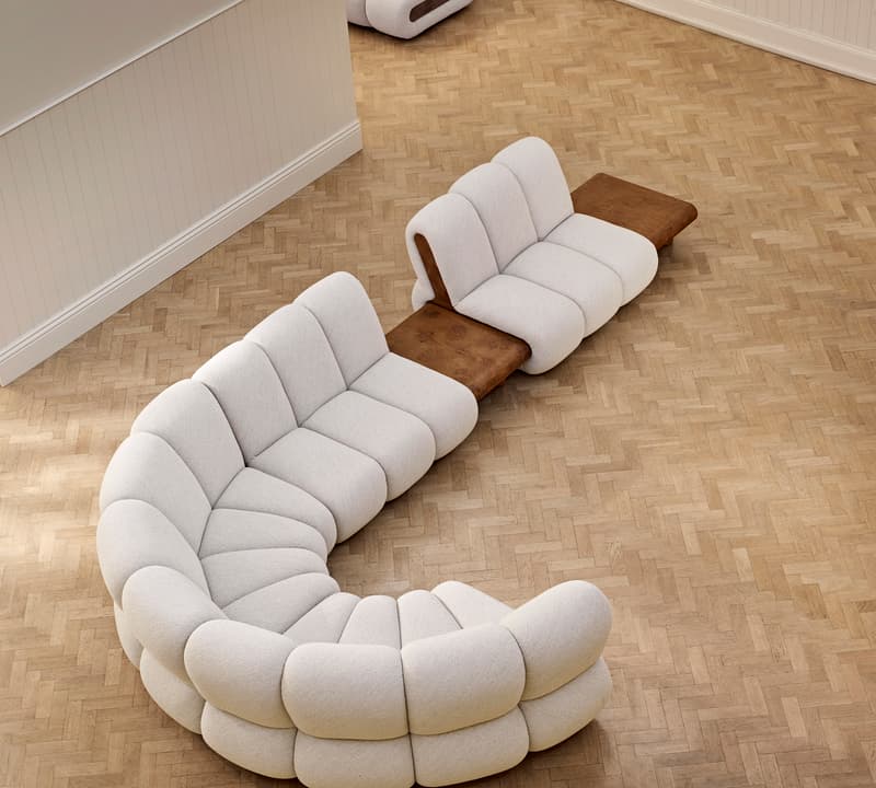 Curved, beige modular sofa with brown wooden side tables placed on a light wooden herringbone floor. Another matching, smaller sofa is located in the back corner of the room.
