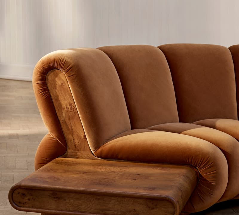 A brown velvet upholstered sofa with a wooden side table attached, placed on a light wooden parquet floor, within a white-paneled room.