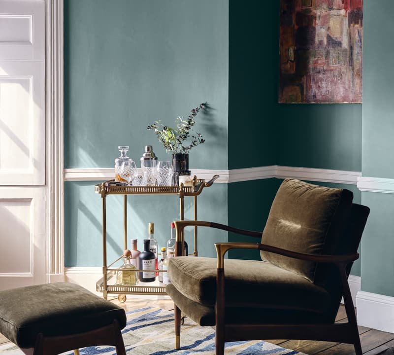 A green armchair with a matching ottoman sits beside a stocked bar cart and a wall-hung abstract painting in a green-walled room with white trim and a patterned rug.