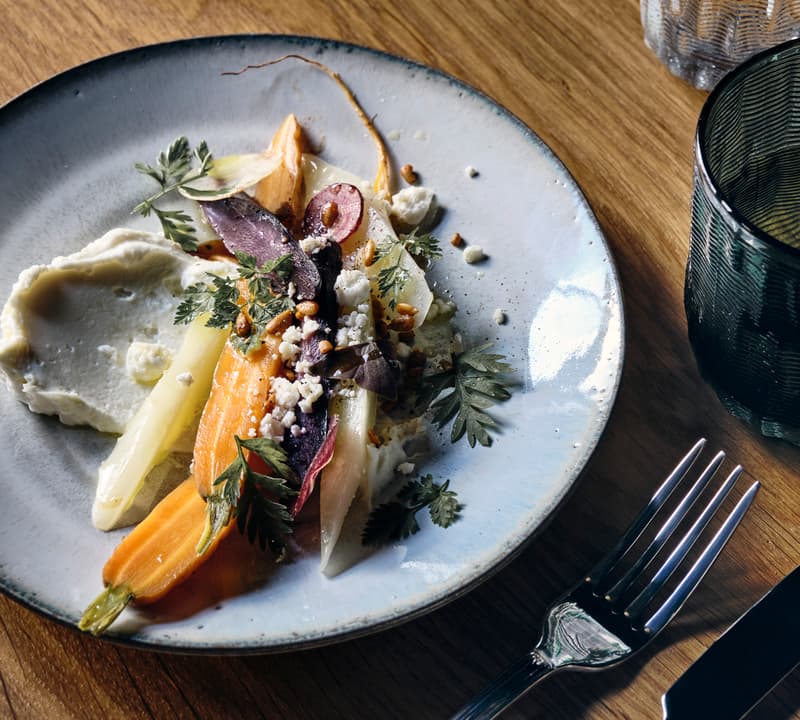 Assorted colorful roasted vegetables garnished with herbs and crumbled cheese on a white plate, accompanied by a fork and knife, resting on a wooden table next to a glass and textured tumbler.