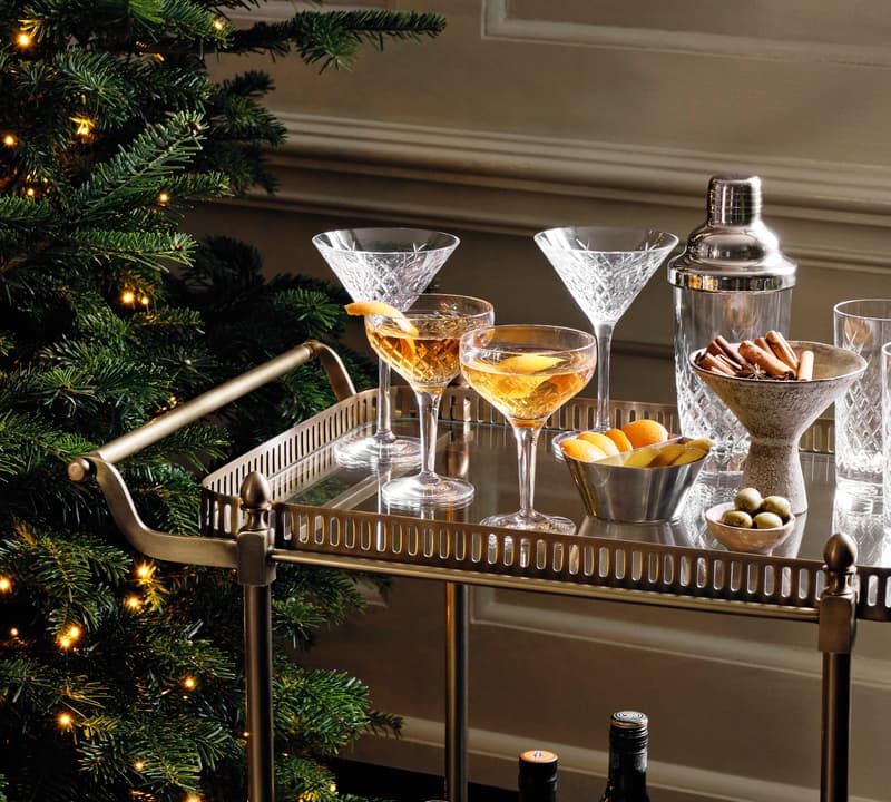 A bar cart with cocktail glasses, a shaker, lemon twists, and olives, stands beside a decorated Christmas tree with string lights in an elegant indoor setting.