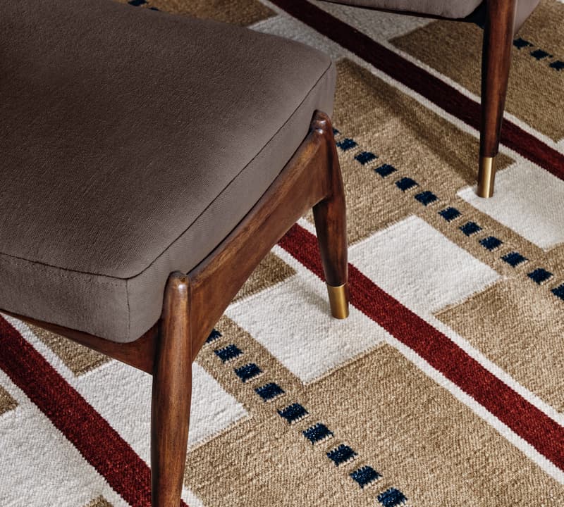 A brown cushioned ottoman with wooden legs and brass tips is situated on a patterned carpet featuring red, white, beige, and blue geometric designs.