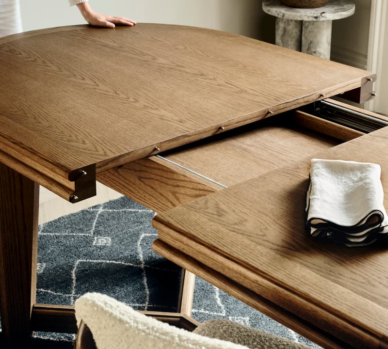 A person extends a wooden dining table in a modern room with a patterned rug, a padded chair, and a plant on a marble side table.