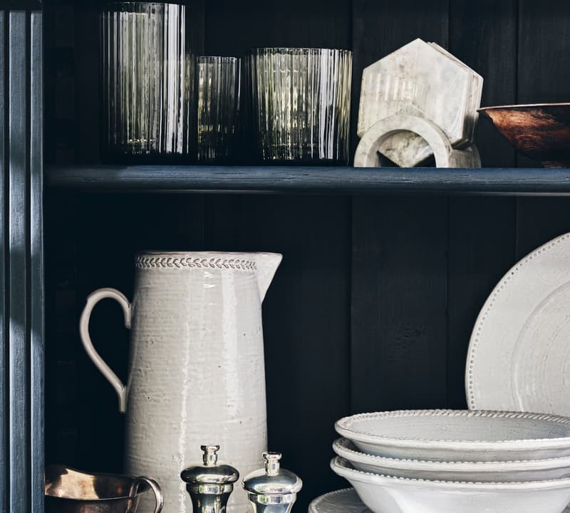 Ceramic pitcher, glass tumblers, metal container, salt and pepper shakers, and stacked white plates rest neatly on dark wooden shelves, creating an organized kitchen display.