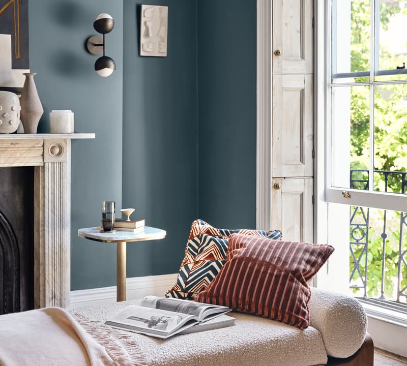 A beige chaise lounge with patterned pillows and an open book sits by a large window in a modern living room with teal walls, a fireplace, and contemporary decor.