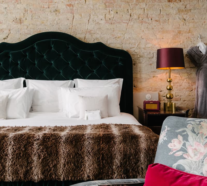 A well-made bed with a dark green velvet headboard and fur blanket sits against an exposed brick wall; beside it, a lamp with a dark red shade and a mannequin. A floral armchair with a red pillow is in the foreground.