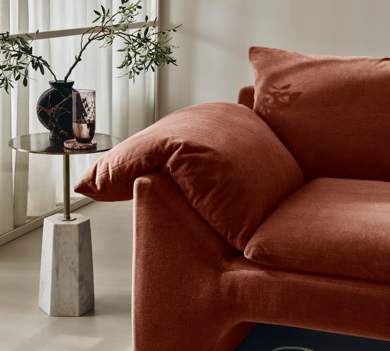 Terracotta sofa is positioned next to a small side table holding a black vase with green branches and a purple glass, set in a modern living room with sheer white curtains.