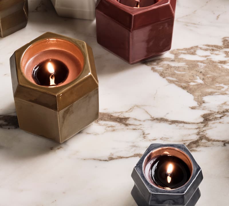 Hexagonal candles with lit wicks are placed on a marble surface. The candles are in ceramic holders of various colors, including gold, red, gray, and cream.