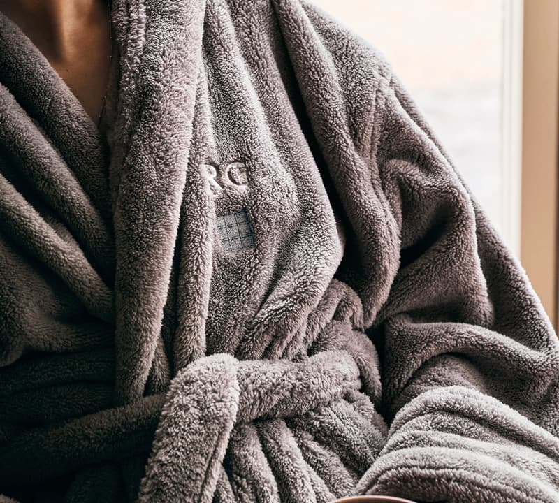 A person wearing a plush gray robe, embroidered with "RCR," holds a cup of coffee while sitting near a window with a blurred outdoor background.