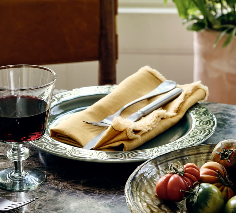 A table holding a wine glass, a plate with a napkin and cutlery, and a bowl of tomatoes in a room with sunlight and a potted plant in the background.